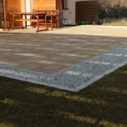 Beżowa kostka brukowa na tarasie z jasnymi kamieniami na obrzeżach, drewniany stół i krzesła w tle, trawnik. Słoneczny dzień.