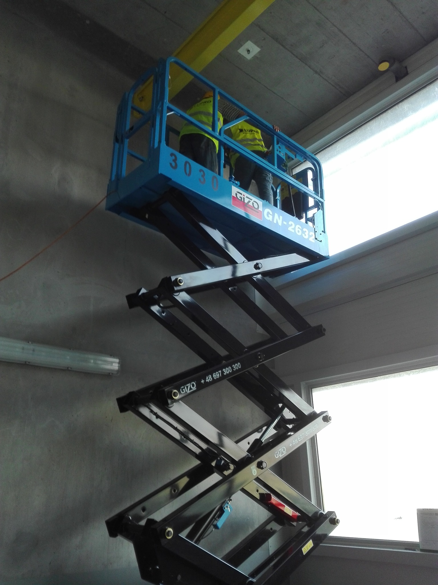 Podnośnik nożycowy z dwoma pracownikami w żółtych kamizelkach, unoszący się blisko sufitu w hali przemysłowej podczas prac montażowych, widoczne elementy konstrukcji hali i okno.