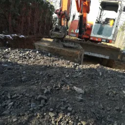 Pomarańczowa koparka Zeppelin w trakcie prac ziemnych, widoczny wykop i gruz. Ślady śniegu na tujach w tle.