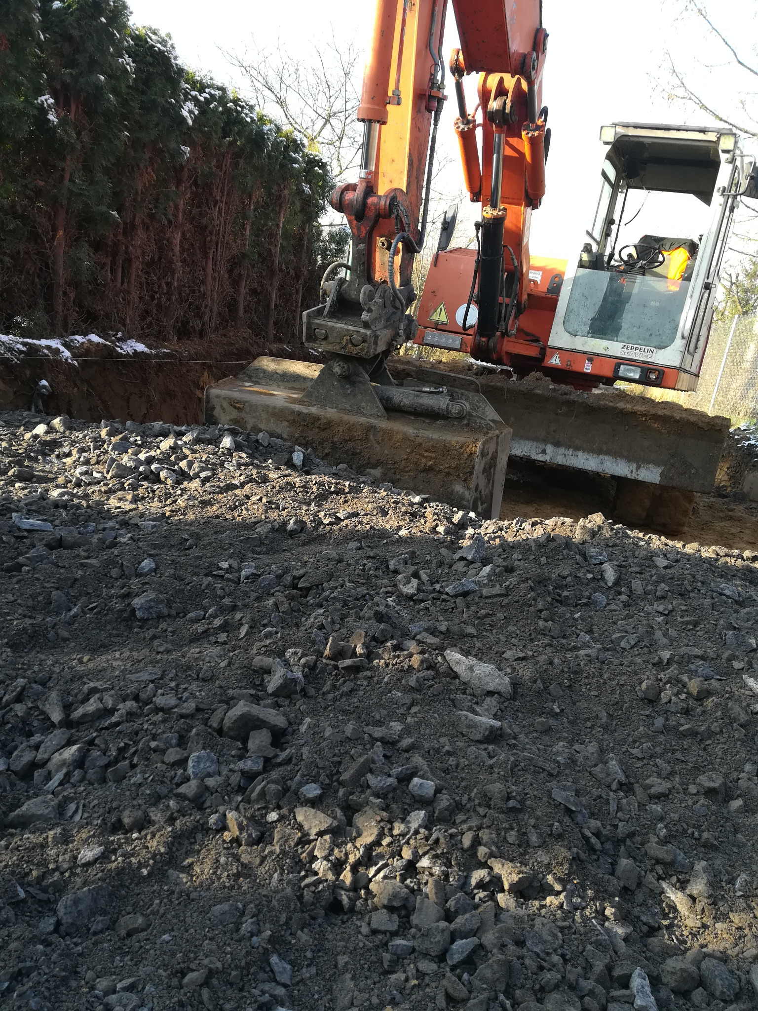 Pomarańczowa koparka Zeppelin w trakcie prac ziemnych, widoczny wykop i gruz. Ślady śniegu na tujach w tle.