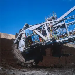 Ogromna koparka kołowa w trakcie pracy, wydobywająca ziemię na tle błękitnego nieba i jasnego wzgórza.