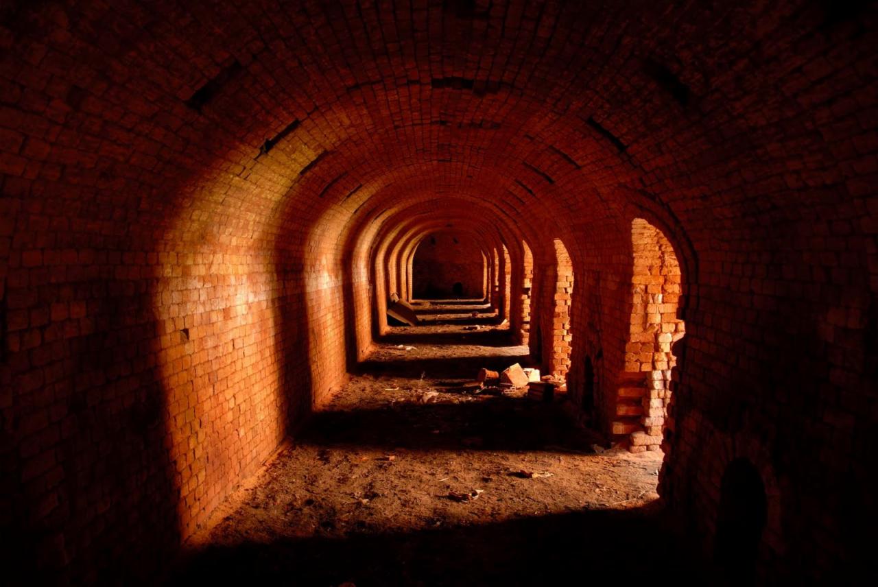 Długi ceglany tunel z łukowymi przejściami, oświetlony ciepłym światłem, ukazujący proces renowacji zabytkowej budowli.