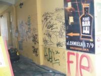 Żółta ściana w przejściu pokryta czarnymi graffiti, obok plakat Piano Bar z czerwoną strzałką i napisami, widoczny fragment podłogi i narożnik ściany.