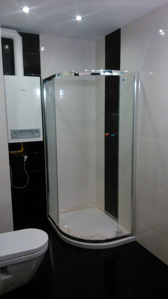 Nowoczesna łazienka z kabiną prysznicową narożną ze srebrnymi profilami, białą ceramiką i czarno-białymi płytkami. Na ścianie zamontowany biały piec gazowy z widoczną instalacją. Minimalistyczny...