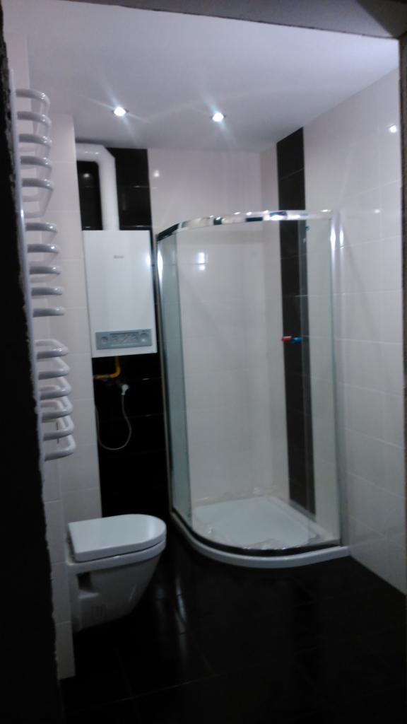 Nowoczesna łazienka z czarno-białymi płytkami, kabiną prysznicową z zaokrąglonymi drzwiami, wiszącą toaletą i grzejnikiem drabinkowym.