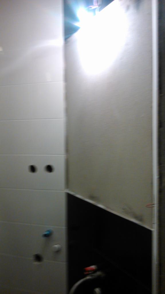 Wnętrze łazienki w trakcie remontu: białe płytki ścienne z wywierconymi otworami, fragment czarnej ściany oraz przygotowana ściana pod dalsze prace wykończeniowe, widoczne rury hydrauliczne.