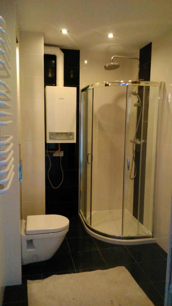 Wykończona łazienka w czarno-białej kolorystyce z kabiną prysznicową z zaokrąglonymi szybami, podwieszaną toaletą, grzejnikiem drabinkowym i piecem gazowym na ścianie.