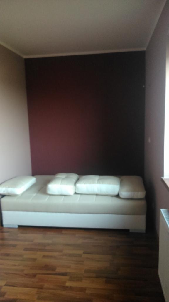 Pokój z brązową ścianą i rozkładaną sofą z jasnymi poduszkami, widoczna drewniana podłoga i grzejnik.