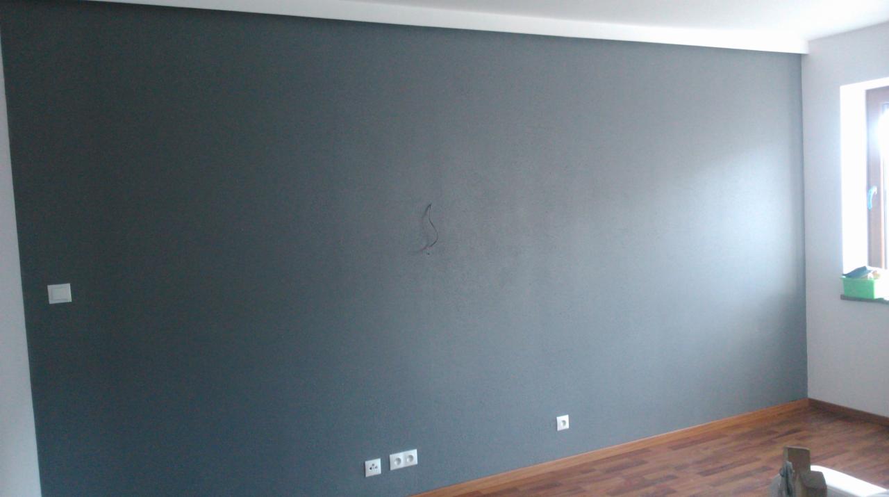 Ściana pomalowana na szaro z widocznymi gniazdkami elektrycznymi, drewniana podłoga i okno z zielonym pudełkiem na parapecie. Na ścianie widoczny ślad po wcześniejszym wierceniu.