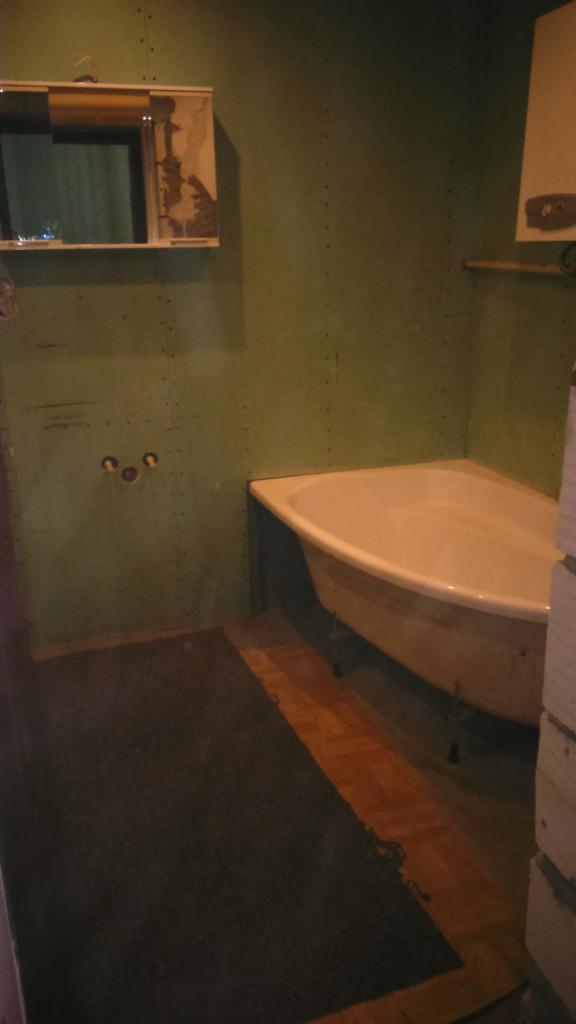 Łazienka w trakcie remontu: widoczna wanna na nóżkach, fragment drewnianej podłogi, ściany pokryte płytami gipsowo-kartonowymi, otwory na instalacje wodne i szafka z lustrem.