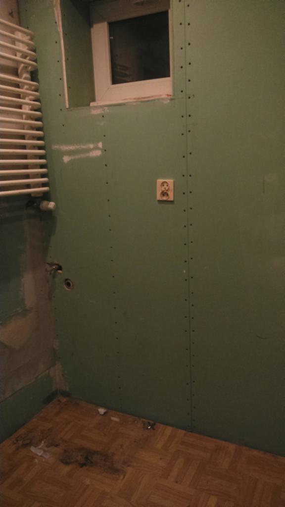 Wnętrze łazienki w trakcie remontu: ściany wyłożone płytami gipsowo-kartonowymi, widoczne łączenia płyt, gniazdko elektryczne, grzejnik drabinkowy i okno. Podłoga wyłożona parkietem. Widoczne ślady...