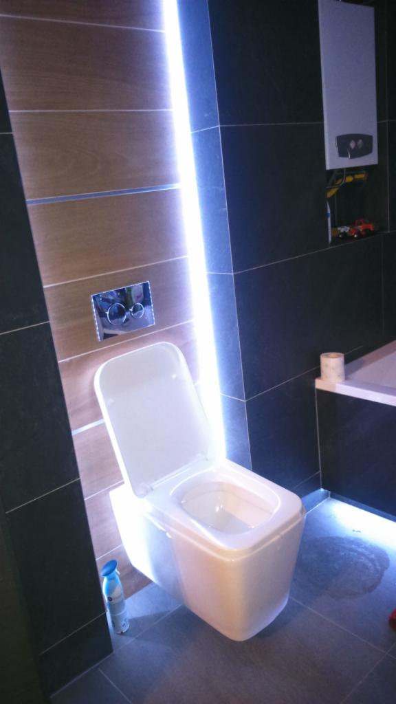 Nowoczesna toaleta wisząca z podświetleniem LED na ścianie i podłogą z ciemnych płytek, widoczny również zbiornik na wodę i odświeżacz powietrza.