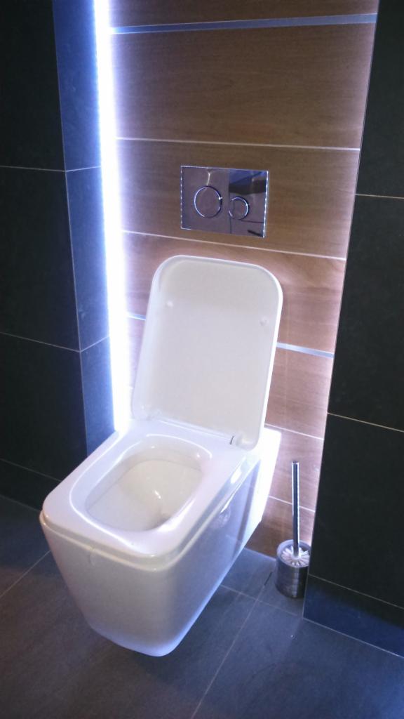 Wisząca toaleta z otwartą klapą, chromowany przycisk spłukiwania na drewnopodobnej ścianie z podświetleniem LED, ciemne płytki podłogowe i ścienne, szczotka do WC w tle.