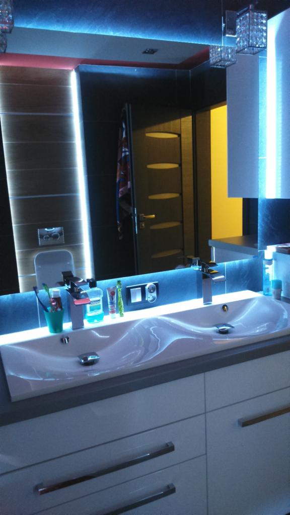 Nowoczesna łazienka z podwójną umywalką o falistym kształcie, podświetlanym lustrem i oświetleniem LED na suficie, odbicie drzwi z trzema owalnymi okienkami w lustrze.