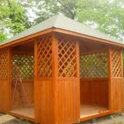 Drewniana altana ogrodowa z kratownicowymi ścianami, widoczne drzewa w tle i drabina oparta o jedną ze ścian.