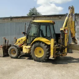 Żółta koparko-ładowarka New Holland na placu budowy, z widocznym łyżką i ramieniem kopiącym, na tle budynku z blachy falistej i błękitnego nieba.