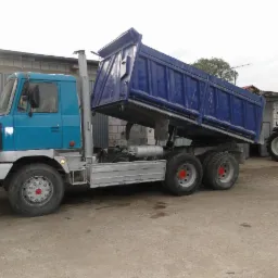 Błękitna ciężarówka wywrotka z podniesioną skrzynią ładunkową na tle budynku z szarej cegły i czerwonego traktora. Widok z boku, lekko z dołu.