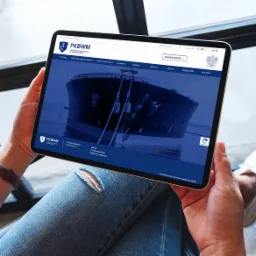 Osoba trzymająca tablet prezentujący stronę internetową Państwowej Komisji Badania Wypadków Morskich (PKBWM) z ilustracją dużego statku na wodzie, widoczne nogi w jeansach z dziurą na kolanie.