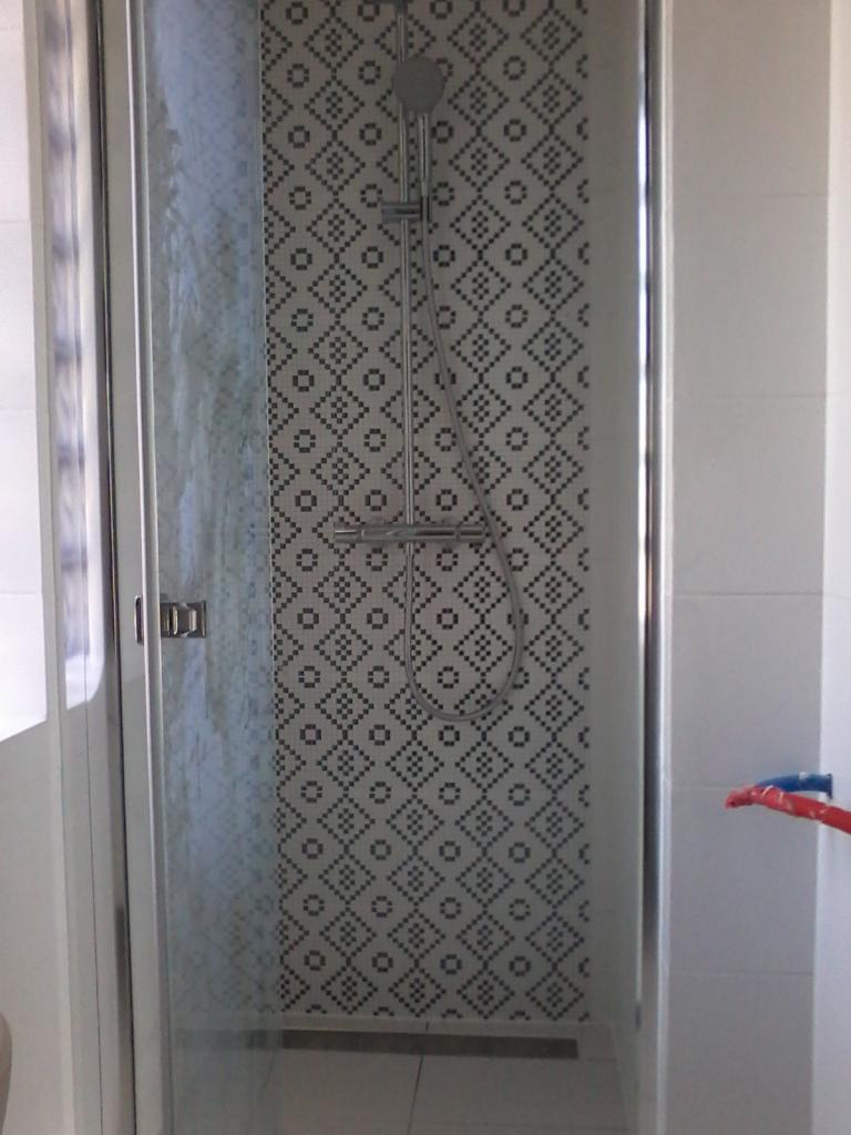 Kabina prysznicowa z wzorzystą mozaiką w geometryczne kształty, chromowana bateria i deszczownica, widoczne fragmenty instalacji wodnej z rurami w kolorze czerwonym i niebieskim.