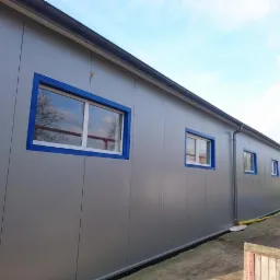 Ściana hali przemysłowej z szarej płyty warstwowej z oknami w niebieskich ramach, widok z zewnątrz.