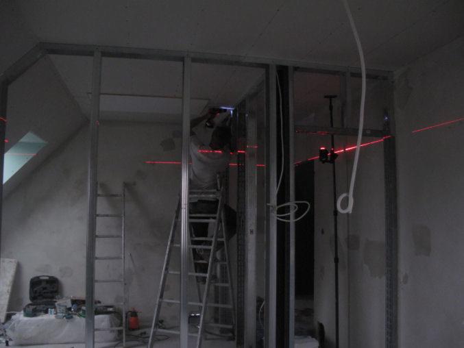 Montaż stelaża z profili stalowych pod zabudowę gipsowo-kartonową na poddaszu, widoczny pracownik na drabinie, laser krzyżowy wyznacza poziom, surowy stan wykończenia.