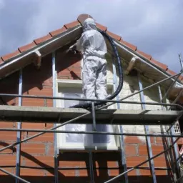 Pracownik w kombinezonie ochronnym aplikuje izolację natryskową pianką PUR na poddasze domu z rusztowania, widoczne cegły i okno zabezpieczone folią.