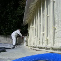 Pracownik w kombinezonie ochronnym aplikuje piankę PUR na elewację budynku, widoczne elementy konstrukcyjne i gruba warstwa izolacji natryskowej, w tle zieleń.