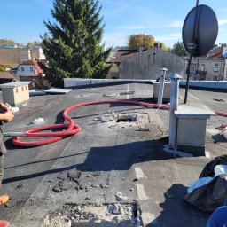 MARGIPS Marcin Biłgorajski - Prace przygotowawcze do ocieplania dachu płaskiego, widoczne otwory, czerwony wąż i kominy wentylacyjne na tle zabudowy miejskiej i wysokiej choinki.