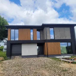 Nowoczesny dom z elewacją z drewnianych paneli i szarego betonu architektonicznego, widok z zewnątrz na etapie wykończenia terenu.