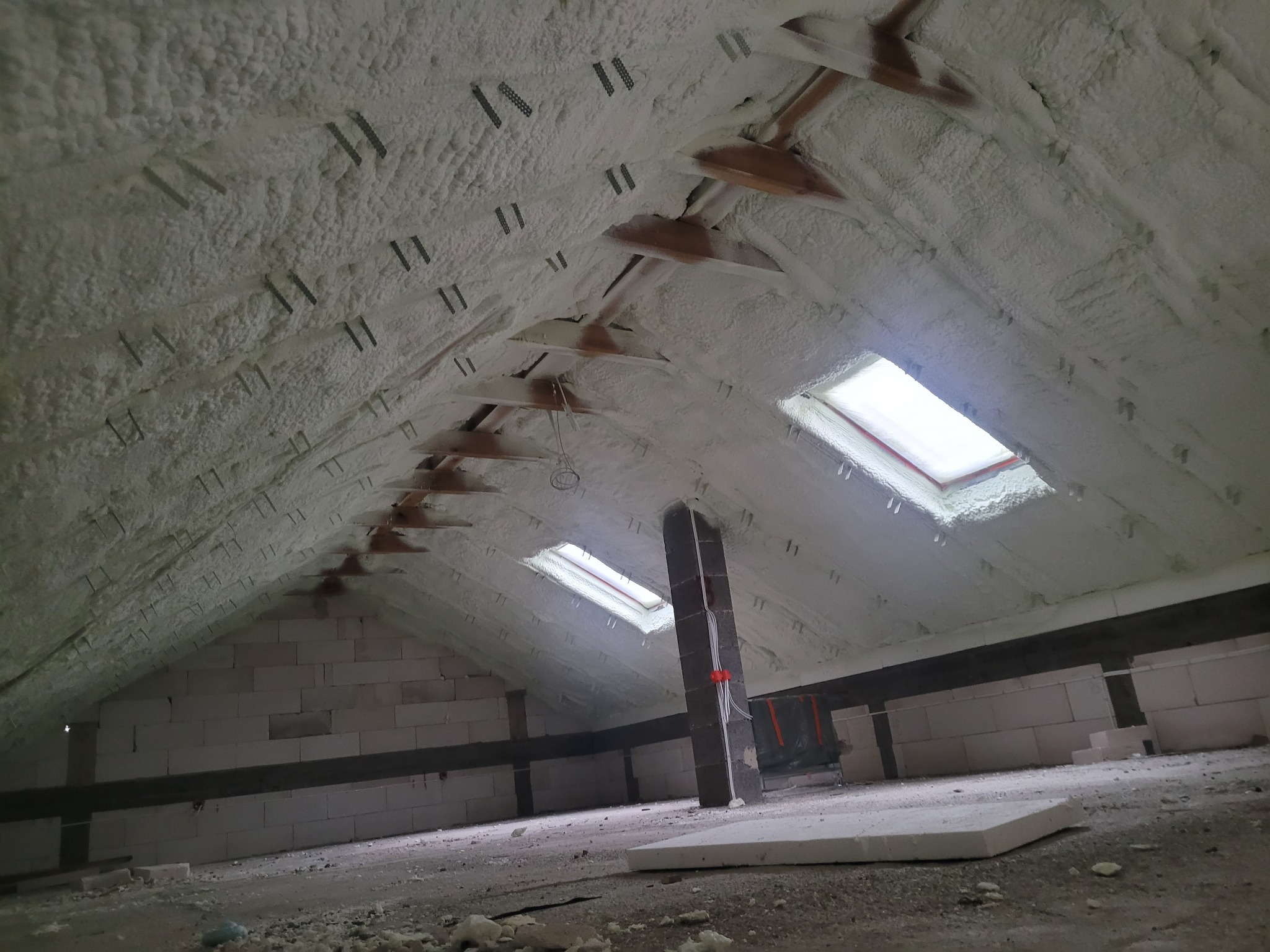 Poddasze po natrysku pianki izolacyjnej, widoczne krokwie, okna dachowe i ściany z bloczków, elementy montażowe w pianie.