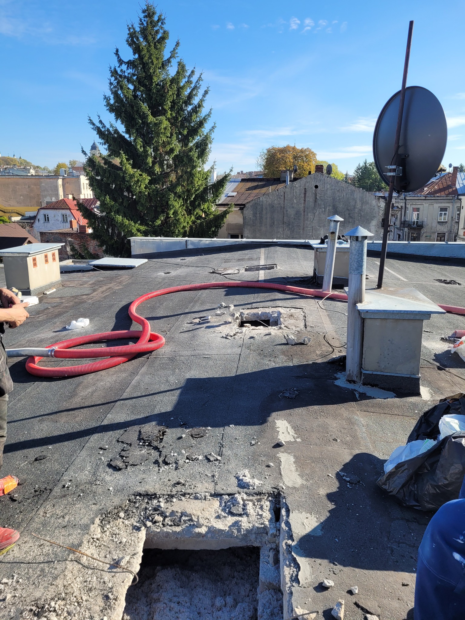 Prace przygotowawcze do ocieplania dachu płaskiego, widoczne otwory, czerwony wąż i kominy wentylacyjne na tle zabudowy miejskiej i wysokiej choinki.