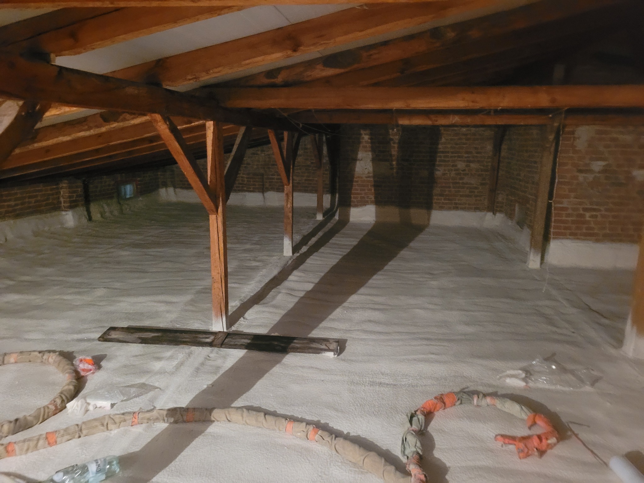 Poddasze z drewnianą konstrukcją pokryte białą pianką izolacyjną, widoczne przewody i butelka wody.