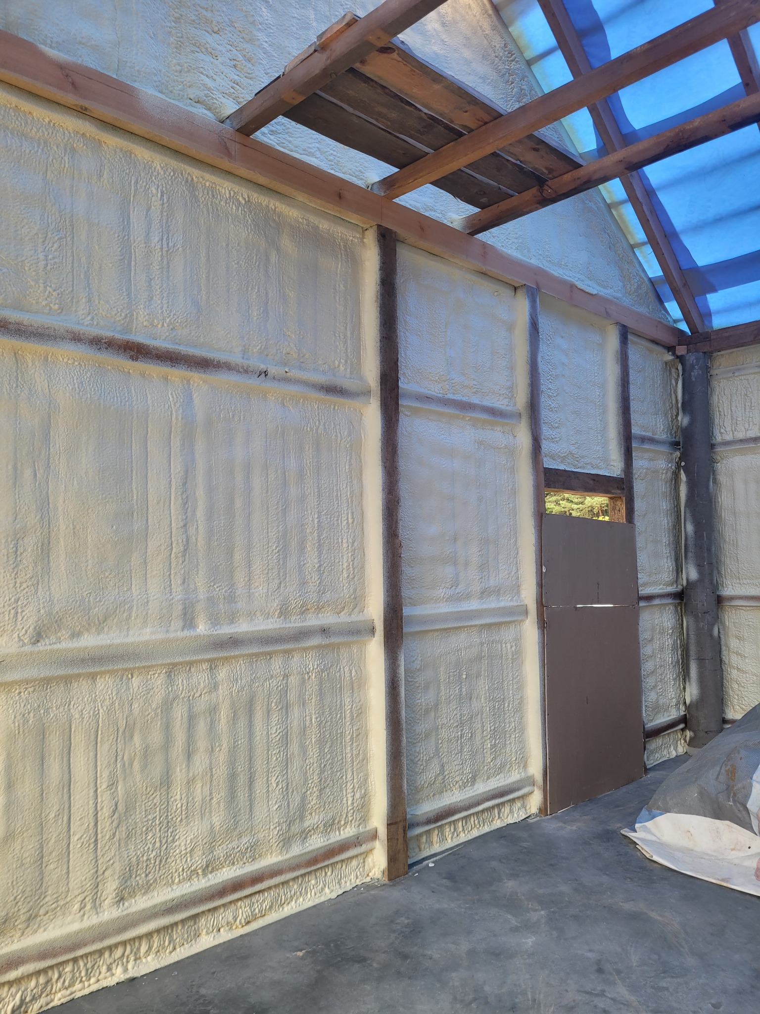 Wnętrze budynku w trakcie ocieplania: ściany i dach pokryte grubą warstwą jasnej pianki izolacyjnej, widoczne drewniane elementy konstrukcyjne i fragment niebieskiej folii. Lekko niedokończone.