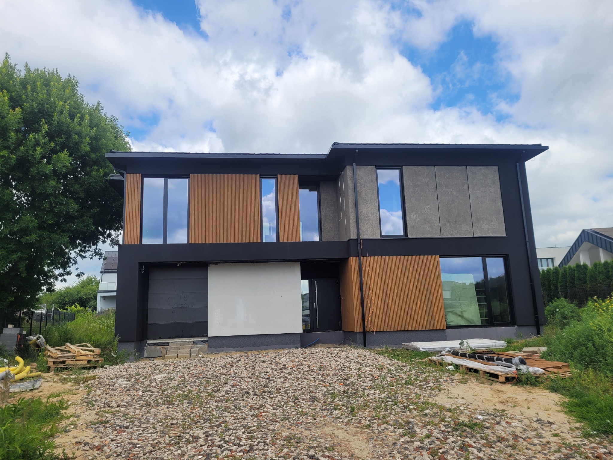 Nowoczesny dom z elewacją z drewnianych paneli i szarego betonu architektonicznego, widok z zewnątrz na etapie wykończenia terenu.