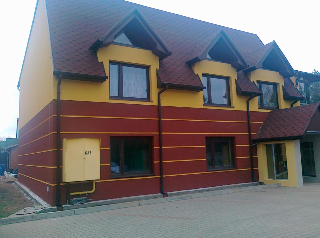 Dwukondygnacyjny budynek z żółto-bordową elewacją, brązowymi oknami i dachem z gontu bitumicznego. Widoczna skrzynka gazowa i kostka brukowa.
