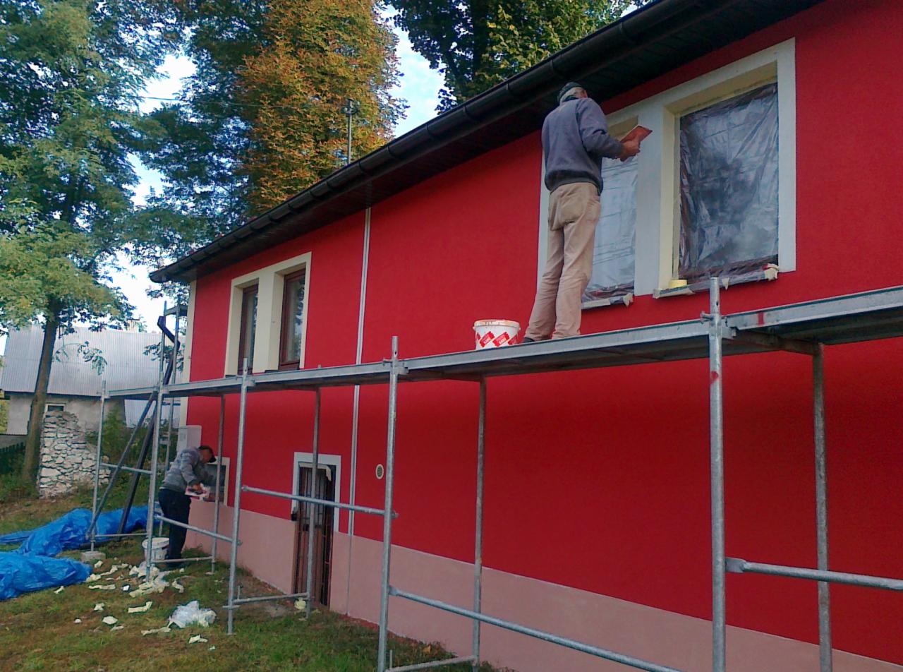 Czerwony dom w trakcie renowacji elewacji. Pracownicy na rusztowaniu szpachlują okna. Część elewacji pomalowana na różowo. Widoczne drzewa i budynki w tle.