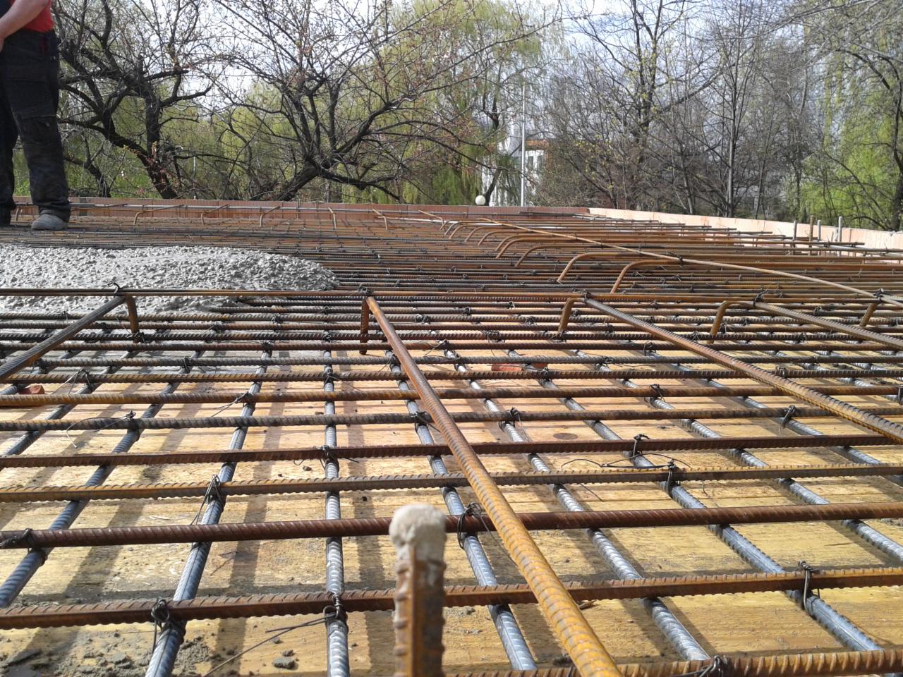 Zbrojenie płyty fundamentowej na deskowaniu, częściowo zalane betonem. Widoczne pręty zbrojeniowe ułożone w siatkę, beton wylewany na zbrojenie, drzewa w tle.