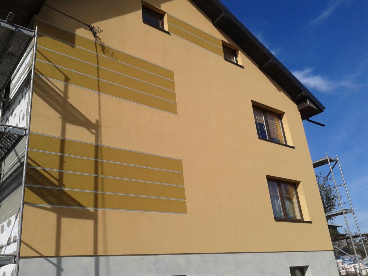 Żółta elewacja domu z dekoracyjnymi pasami w odcieniach brązu, widoczna z rusztowaniem po lewej stronie, na tle błękitnego nieba. Detal wykończenia zewnętrznego.