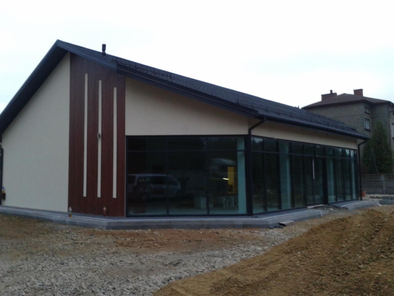 Nowoczesny budynek usługowy z dużymi przeszkleniami i elewacją w odcieniach beżu i brązu, widoczny na tle pochmurnego nieba i sąsiedniego budynku.