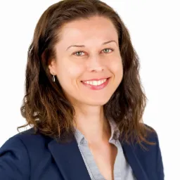 Portret uśmiechniętej kobiety w granatowej marynarce i szarej koszuli, z kolczykiem w kształcie piramidy.