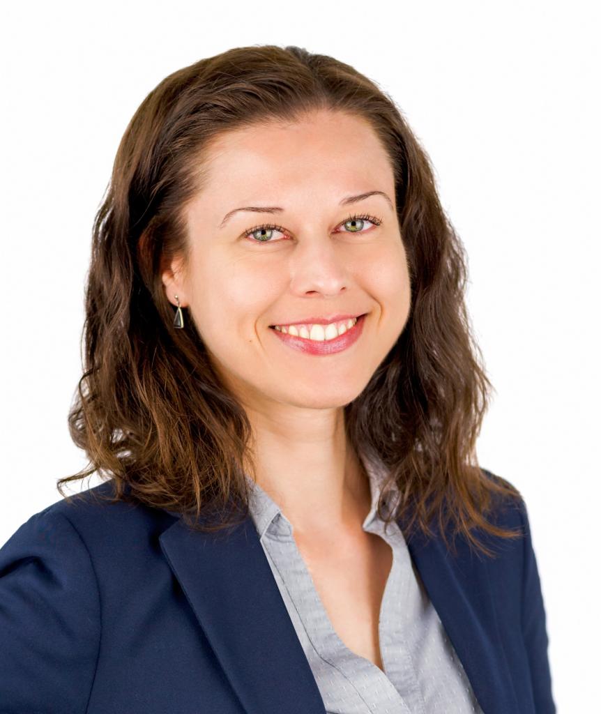 Portret uśmiechniętej kobiety w granatowej marynarce i szarej koszuli, z kolczykiem w kształcie piramidy.