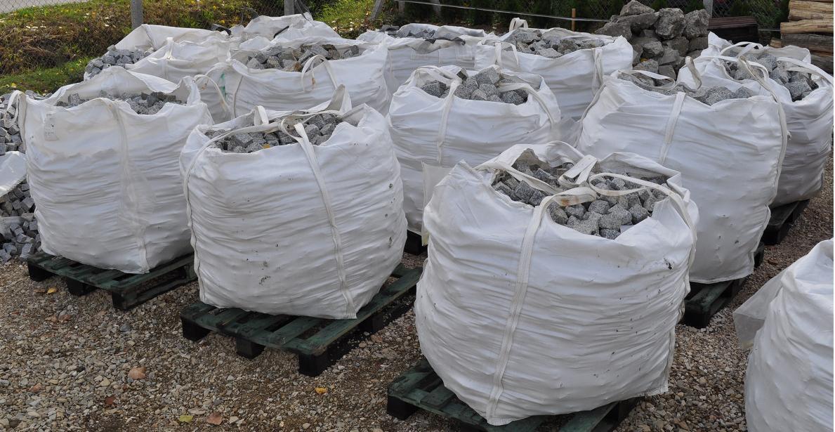 Białe worki typu big bag wypełnione kostką granitową Strzegom, ułożone na zielonych paletach, na żwirowym podłożu, w tle ogrodzenie z siatki.