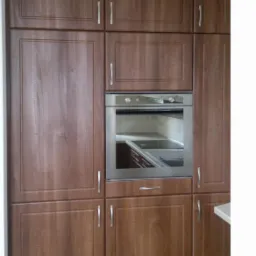 Zabudowa kuchenna z drewnianymi frontami, srebrne uchwyty, wbudowany piekarnik ze stali nierdzewnej, oświetlenie punktowe na suficie.