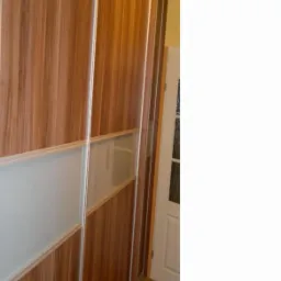 Wysoka szafa przesuwna z drewnopodobnym fornirem i matowymi szklanymi wstawkami, widoczna z boku, z drzwiami prowadzącymi do innego pomieszczenia w tle.