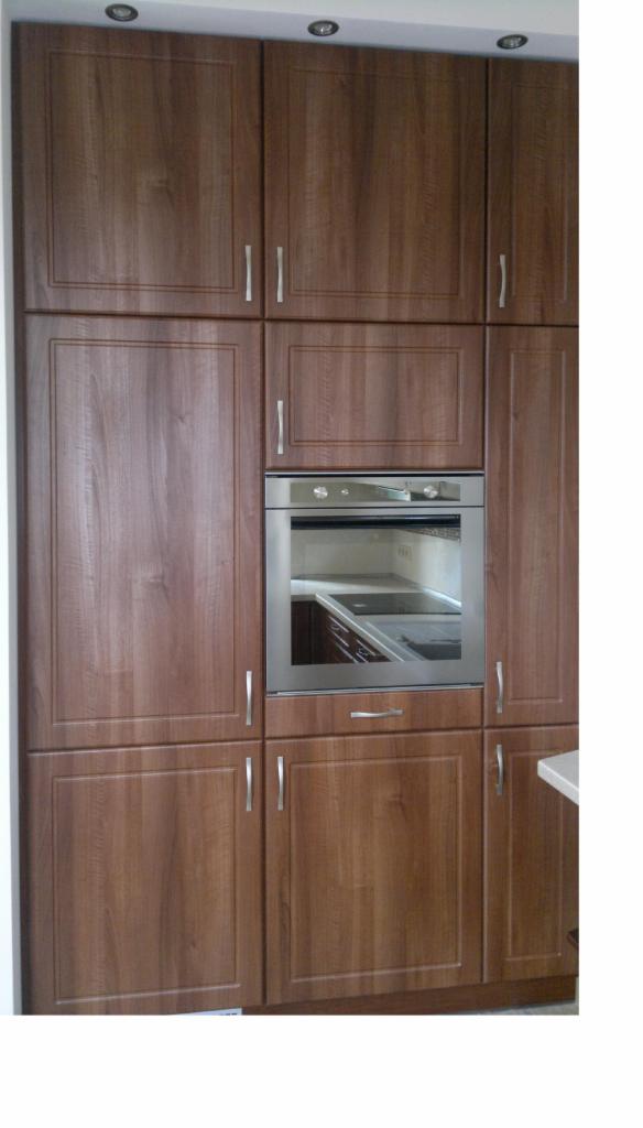 Zabudowa kuchenna z drewnianymi frontami, srebrne uchwyty, wbudowany piekarnik ze stali nierdzewnej, oświetlenie punktowe na suficie.