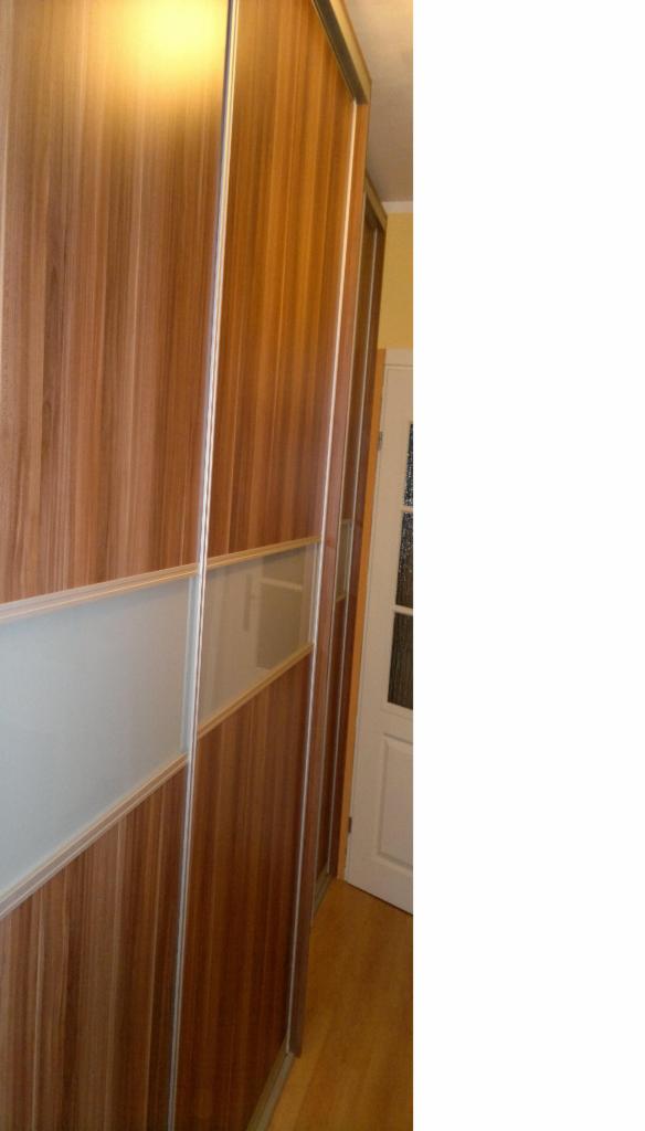 Wysoka szafa przesuwna z drewnopodobnym fornirem i matowymi szklanymi wstawkami, widoczna z boku, z drzwiami prowadzącymi do innego pomieszczenia w tle.