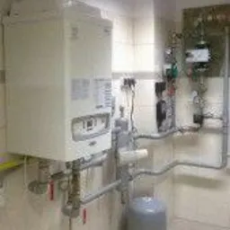 Biały piec CO zamontowany na ścianie w pomieszczeniu z jasnymi płytkami. Widoczne rury, zawory i zbiornik wyrównawczy. Oświetlenie sufitowe. Ujęcie z dołu.