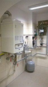 Biały piec CO zamontowany na ścianie w pomieszczeniu z jasnymi płytkami. Widoczne rury, zawory i zbiornik wyrównawczy. Oświetlenie sufitowe. Ujęcie z dołu.