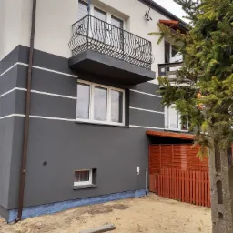 Dwukondygnacyjny dom z szarą elewacją, białymi pasami dekoracyjnymi, balkonem z kutą balustradą i brązową rynną, widok z boku na niezagospodarowaną działkę z piaskiem.