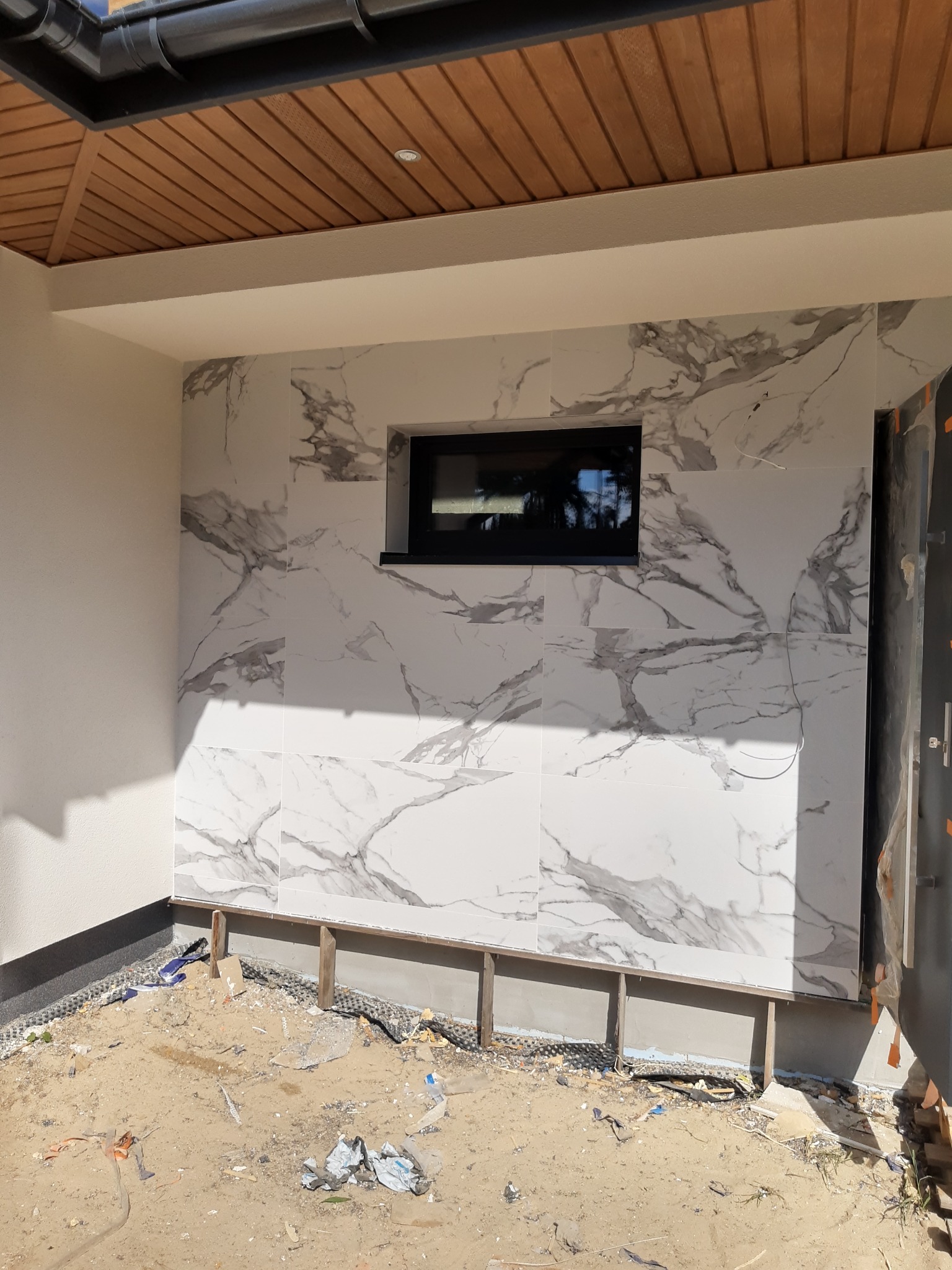 Ściana budynku w Dreństwie wykończona białymi płytkami imitującymi marmur, z czarnym oknem, widoczne przygotowania do dalszych prac wykończeniowych.